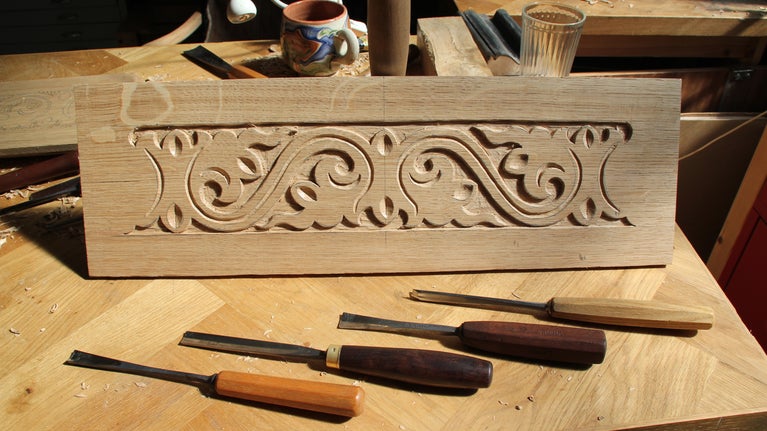 A pattern with swirls and waves carved into a piece of wood, with carving tools laying on top of the piece of wood.